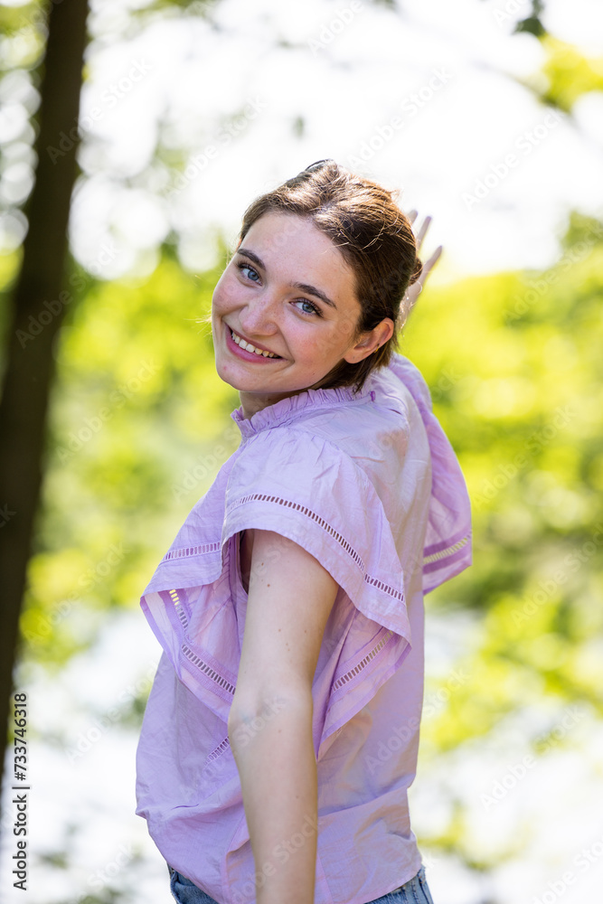 This image shows a young woman turning to smile over her shoulder, her eyes gleaming with playfulness and joy. She is outside on a sunny day, with trees providing a dappled light backdrop. Her light This image shows a young woman turning to smile over her shoulder, her eyes gleaming with playfulness and joy. She is outside on a sunny day, with trees providing a dappled light backdrop. Her light
