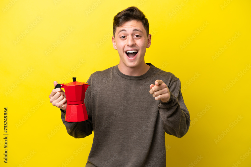Young Brazilian man holding coffee pot isolated on yellow background surprised and pointing front Young Brazilian man holding coffee pot isolated on yellow background surprised and pointing front