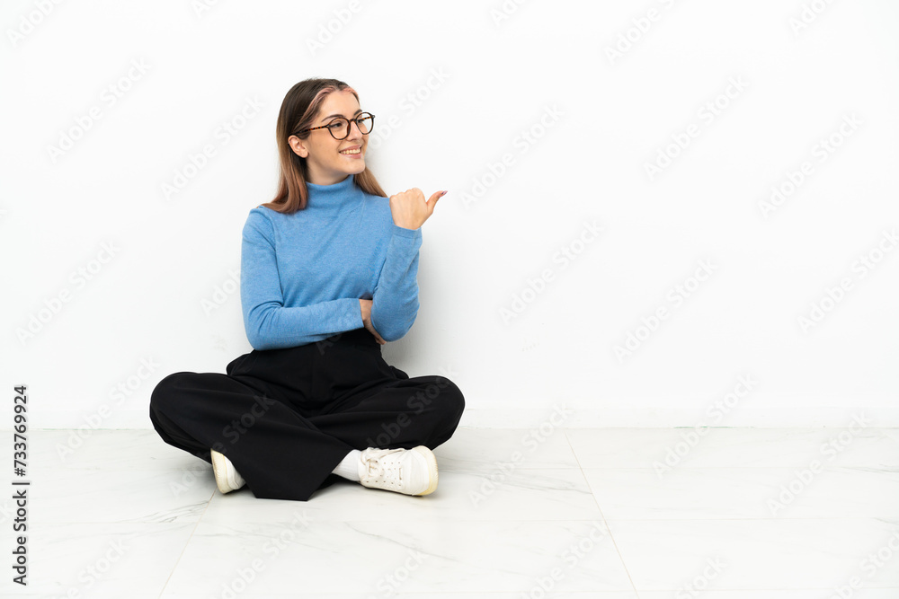 Young Caucasian woman sitting on the floor pointing to the side to present a product Young Caucasian woman sitting on the floor pointing to the side to present a product