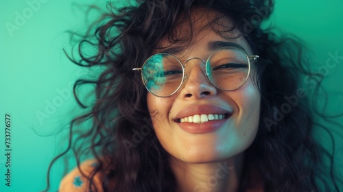 A young woman with curly hair wearing round glasses smiling with her eyes closed against a teal background.