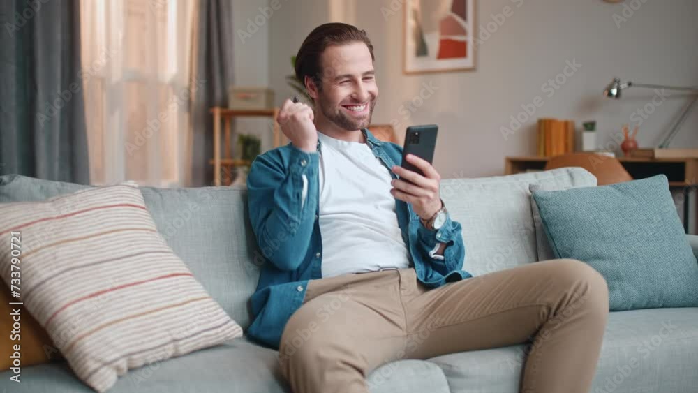 Cheerful man sitting on sofa and using smartphone. Male sitting on couch and shopping distantly. Happy man successfully finishing internet purchase. Male holding credit card and rejoicing. Cheerful man sitting on sofa and using smartphone. Male sitting on couch and shopping distantly. Happy man successfully finishing internet purchase. Male holding credit card and rejoicing.