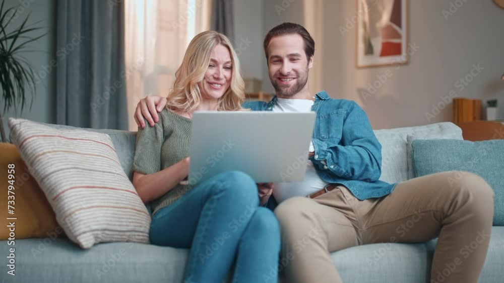 Young couple sitting on couch and talking actively. Man hugging woman by shoulder. Male and female choosing movie. Man and woman relaxing at home. People in love holding laptop. Young couple sitting on couch and talking actively. Man hugging woman by shoulder. Male and female choosing movie. Man and woman relaxing at home. People in love holding laptop.