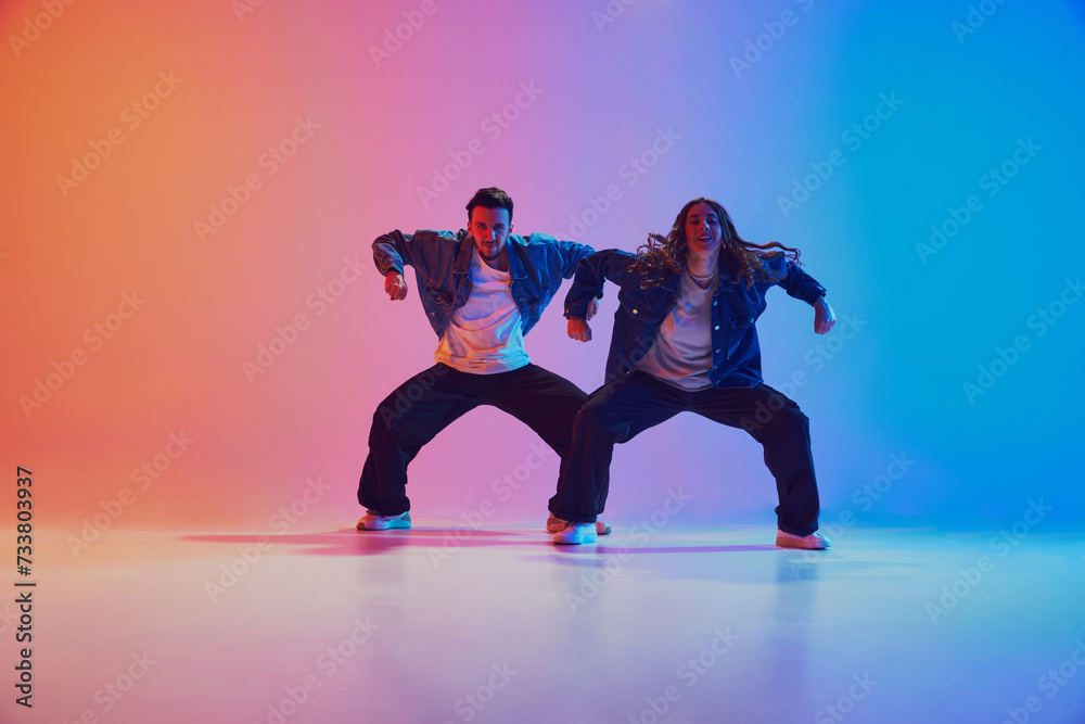 Energetic dance pair in denim jackets in squatting dance pose in neon-lit studio against gradient blue-orange background. Concept of youth culture, music, lifestyle, style and fashion, action.