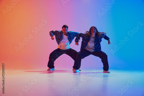 Fototapeta Naklejka Na Ścianę i Meble -  Energetic dance pair in denim jackets in squatting dance pose in neon-lit studio against gradient blue-orange background. Concept of youth culture, music, lifestyle, style and fashion, action.