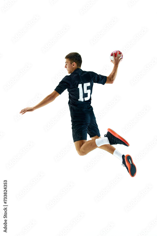Full-length side view image of young man in dark uniform, handball player in motion, in a jump with ball during game, training over white studio background. Concept of sport, tournament, competition