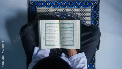 Top view of a Muslim man reading the Quran on a prayer mat while holding prayer beads.