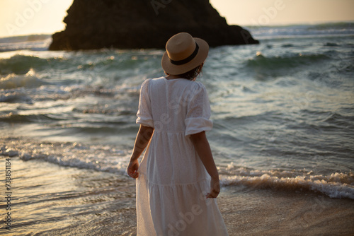 a young girl in a white dress walks on the beautiful ocean shore with sunset light and beautiful sea waves