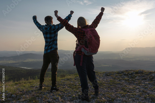Two young hikers, a guy and a girl, walking uphill to the mountain top, celebrating success, raising their arms. Happiness, challenge, goal, and achievement concepts.