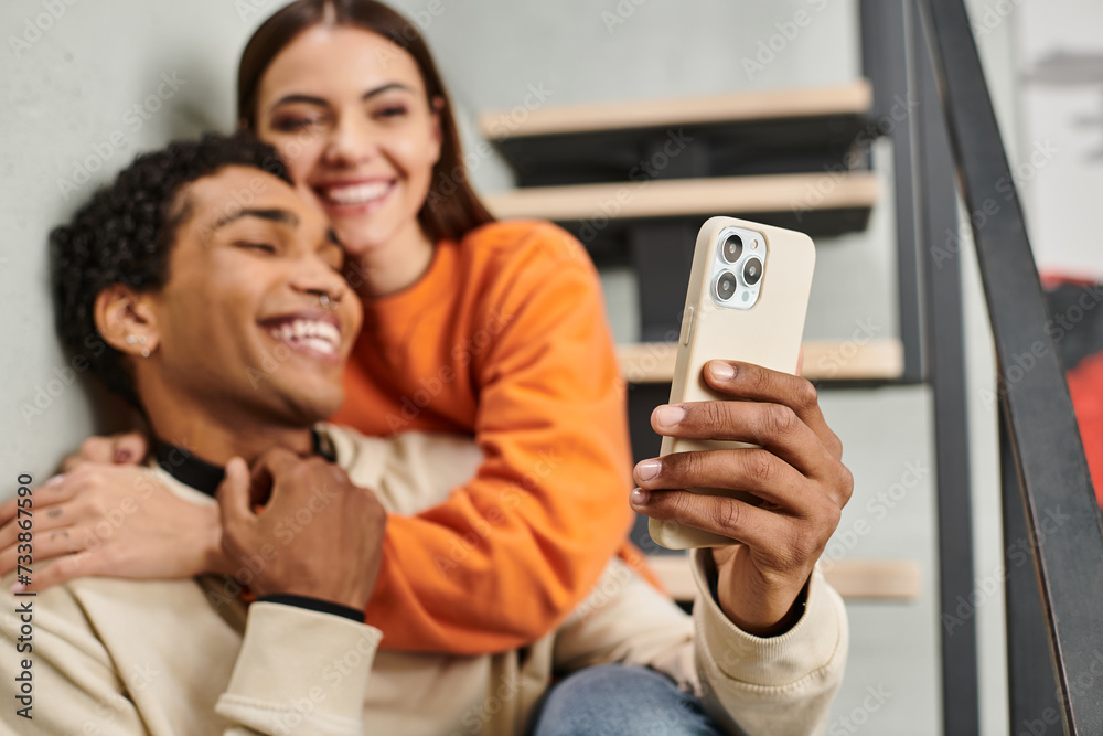 cheerful diverse couple sitting on stairs and smiling while taking selfie on smartphone in hostel cheerful diverse couple sitting on stairs and smiling while taking selfie on smartphone in hostel