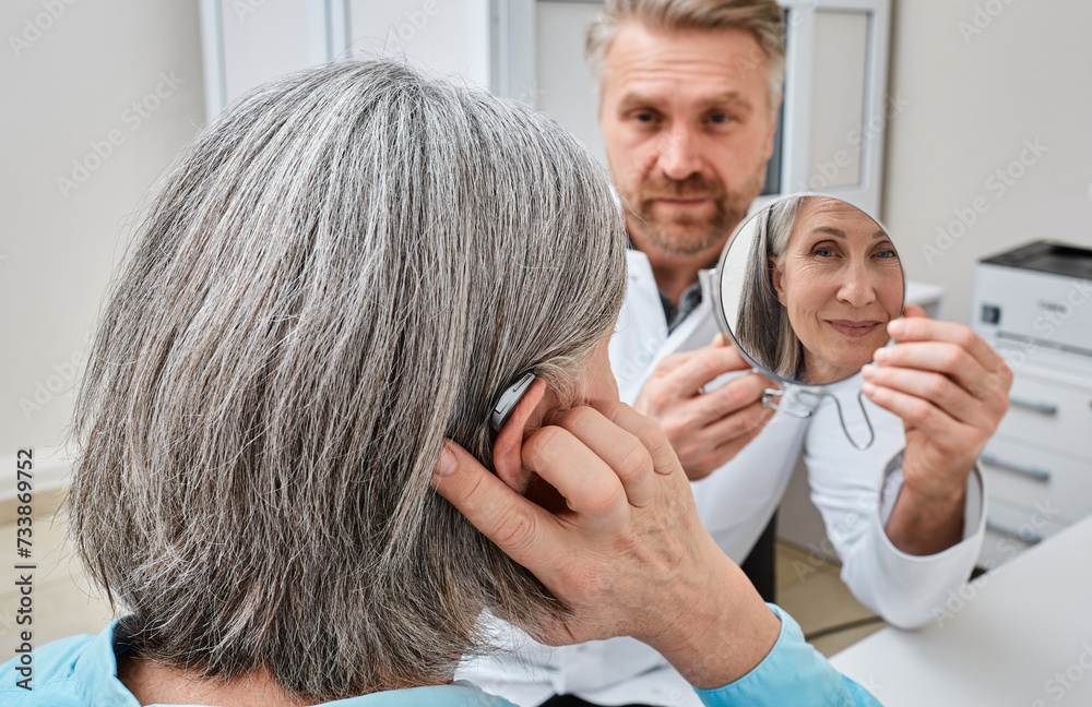 Senior woman trying on a hearing aid while looking into a mirror held by a hearing aid specialist in audiology clinic. Stylish hearing device Senior woman trying on a hearing aid while looking into a mirror held by a hearing aid specialist in audiology clinic. Stylish hearing device