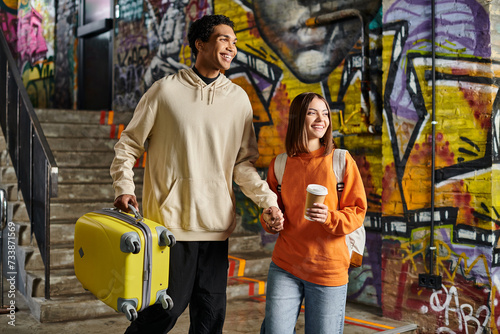 Cheerful couple holding hands and walking with a yellow suitcase in a graffiti-painted wall, hostel