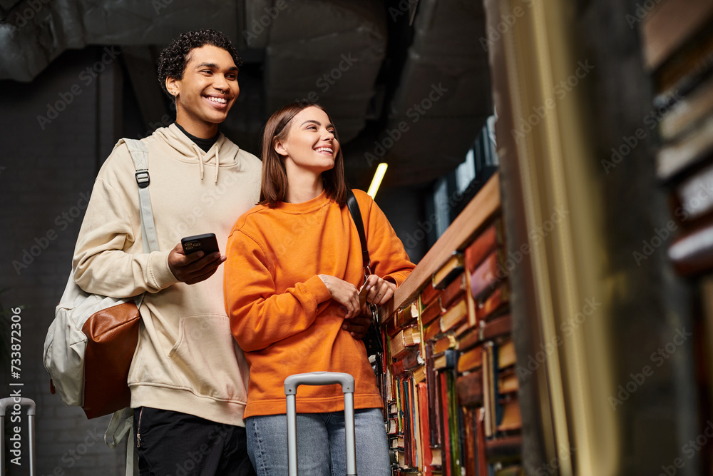 happy and diverse couple with smartphone sharing a joyful moment in hostel near a bookshelf