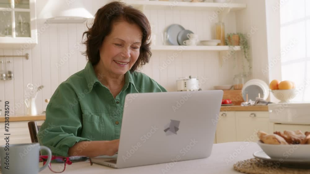 elderly woman sits at table in kitchen in morning with laptop. types on keyboard looks intently in screen, smiles. succeed in using computer and internet, feels pleased and comfortable elderly woman sits at table in kitchen in morning with laptop. types on keyboard looks intently in screen, smiles. succeed in using computer and internet, feels pleased and comfortable