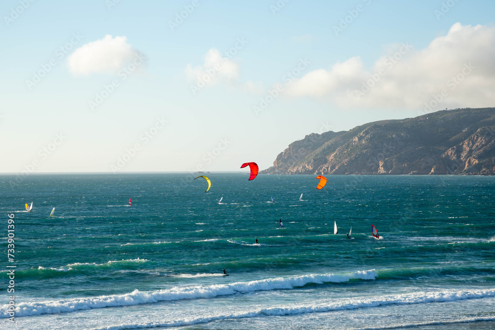 View of the Atlantic Ocean with surfers and windsurfers in the water against of the blue sky and mountains. A beautiful landscape with a view of the ocean. Praia do Guin, Cascais. Portugal.