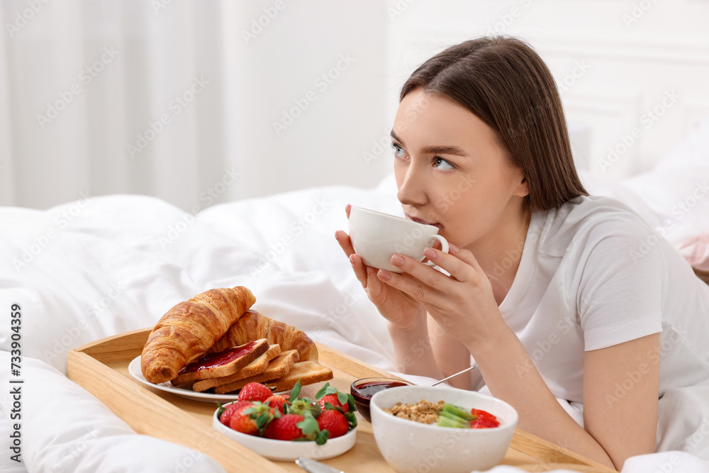 Beautiful woman drinking coffee near tray with breakfast on bed. Space for text