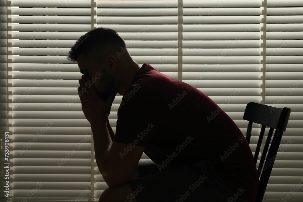 Sad man sitting near closed blinds indoors