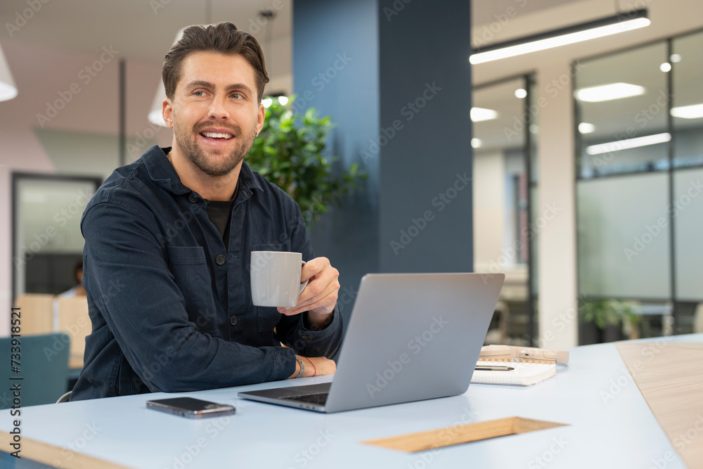 Smiling businessman working on laptop and drinking coffee in modern office Smiling businessman working on laptop and drinking coffee in modern office