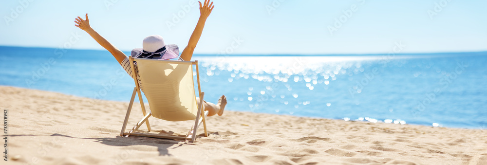 Happy woman sitting with raised hands in the chaise-lounge on the sea beach. - obrazy, fototapety, plakaty 
