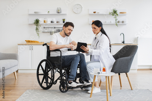 Full length shot of female brunette doctor checking up condition of bearded male patient at home. Nurse talking to mature man on wheelchair, showing examination results using modern tablet. photo