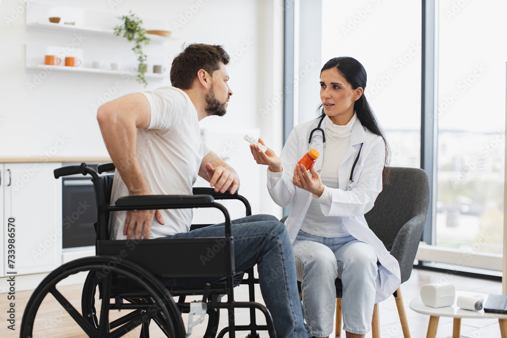 Female general practitioner communicating with patient with disability holding bottle of medication. Doctor discussing health issues with caucasian male with pain in lower back of vertebral discs.