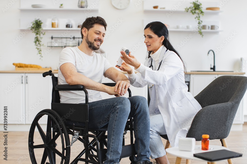 Portrait of pleasant brunette 30-aged woman doctor, visiting her mature male patient with disability at home, showing him results of body temperature measurement on digital infrared thermometer.