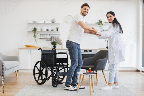 Medical specialist supporting patient in living room of home. Full length view of attractive Caucasian woman helping young patient with disability to stand from wheelchair in modern light kitchen.