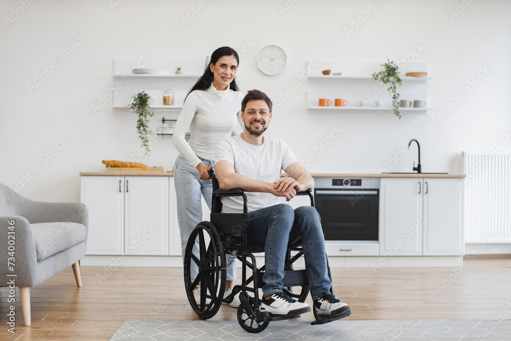 Full length portrait of Caucasian wheelchair user and his wife smiling at camera while resting in open-plan kitchen of apartment. Cheerful young family in casual clothes spending pastime indoors.