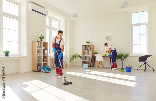 Young male cleaners in overalls, performing room cleaning duties, professional male team busy sanitizing, sweeping, polishing, mopping floor, house cleaning services for home, office