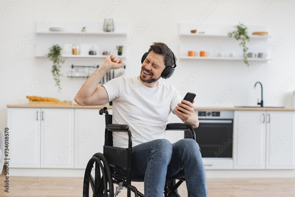 Cheerful Caucasian adult dancing along while hearing loud tunes in headphones. Joyous man in white t-shirt raising hand while looking at smartphone sitting in wheelchair in studio apartment. Cheerful Caucasian adult dancing along while hearing loud tunes in headphones. Joyous man in white t-shirt raising hand while looking at smartphone sitting in wheelchair in studio apartment.