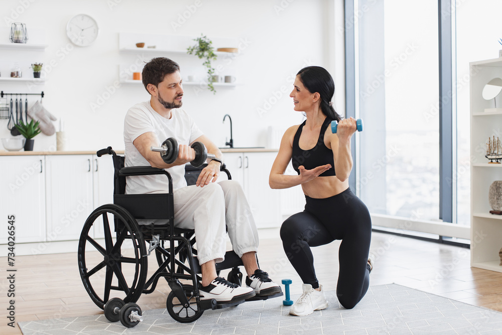 Obraz premium Rehab fitness for young male patient. Millennial physiotherapist in sportswear helping adult man with a disability in wheelchair to workout with dumbbells, to recover from injury at home.