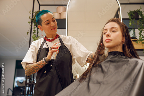 Stylish female hairdresser applying spray on wet hair to woman in beauty salon