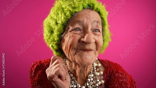Fisheye POV of funny mature elderly woman, 80s, having video chat with friends and family in front of computer, wearing green hat isolated on pink background.