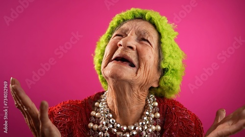 Slow motion view of concerned, unhappy elderly senior old funny crazy woman with wrinkled skin green wig or hat and no teeth put hands out ask WHY ME, isolated on pink background studio.