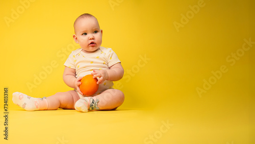 Beautiful baby girl holds orange sitting in studio on yellow background.