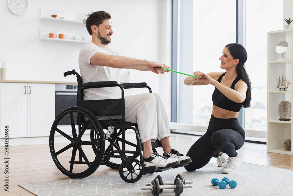 Obraz premium Professional physiotherapist in sportswear helping adult man with a disability in wheelchair to workout with stretching bands, to recover from injury at home. Rehab fitness for young male patient.