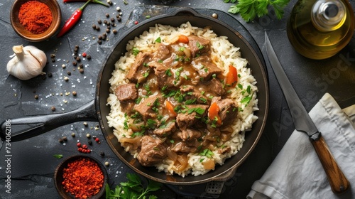 The image displays a savory beef stew with tender chunks of meat and finely chopped vegetables, like carrots, served atop a bed of fluffy white rice, all contained in a cast-iron skillet. The skillet 