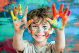 Smiling boy playing with colors, paint on hands and face, showing the two hands full of colorful