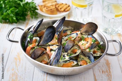 Mussels clams in cream sauce in cooking pan and toasted bread on wooden table.