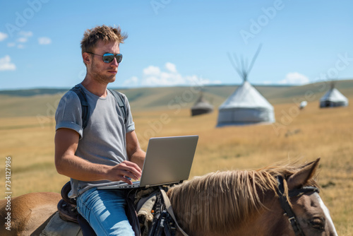 A young digital nomad wearing sunglasses sits on a horse with his laptop in a field, traditional yurts visible in the background under a clear blue sky. photo