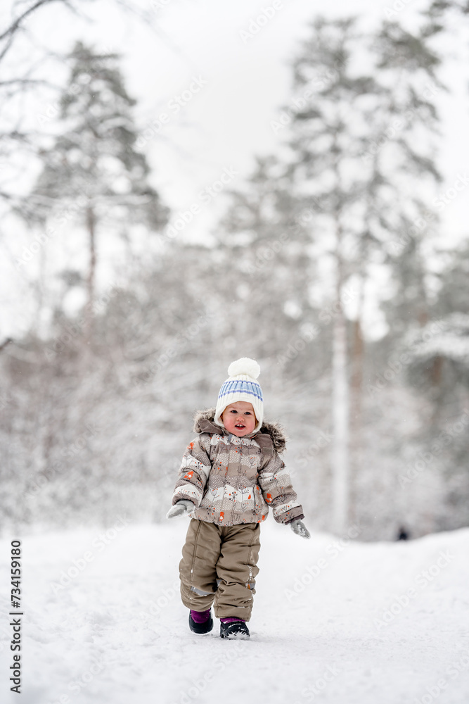 Fototapeta premium Little baby child in a warm jumpsuit walks in a snowy winter forest