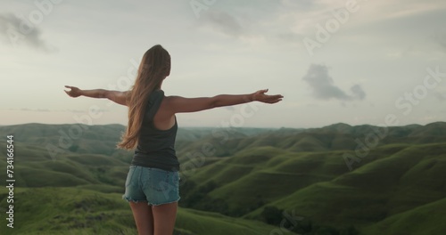 Woman enjoy sunset landscape, raises hands against green hills valley. Long hair brunette girl looking on beautiful nature scene. Freedom, happiness lifestyle. Travel, tourism, holiday.