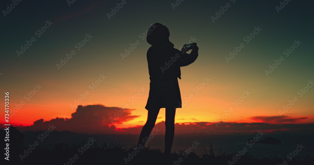 Girl silhouette captures sunset sky nature beauty. Female make photo on her phone standing over sea, enjoy bright colorful sky. Outdoor lifestyle travel, summer holiday vacation. Back view