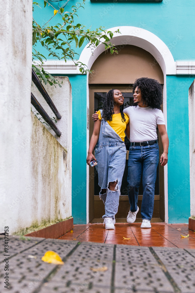 Latinx companions, one with a large afro, enjoying a friendly chat by a blue house entrance.