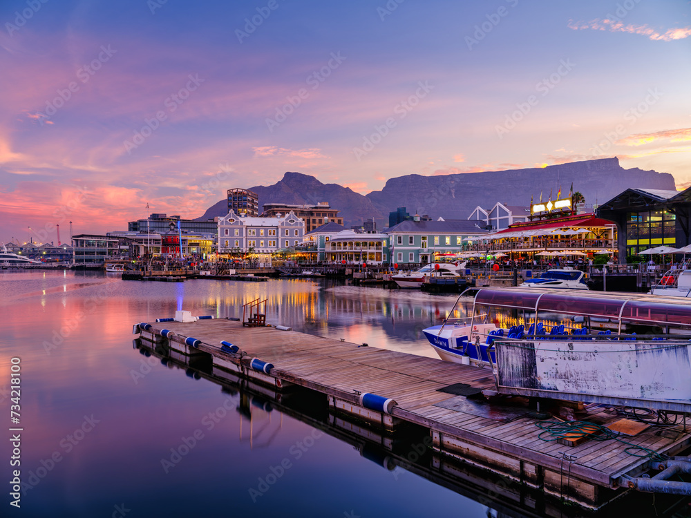 Obraz premium Victoria and Alfred, V and A waterfront lit up during a colorful sunset with table mountain in the background, Cape Town, South Africa