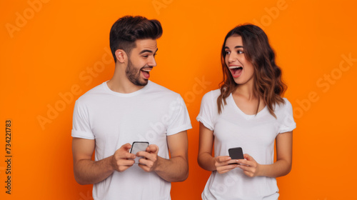 young man and woman are both holding smartphones and smiling at each other, with a bright orange background