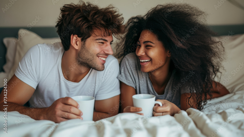 happy couple is lying in bed, holding white mugs, and sharing a joyful moment together.