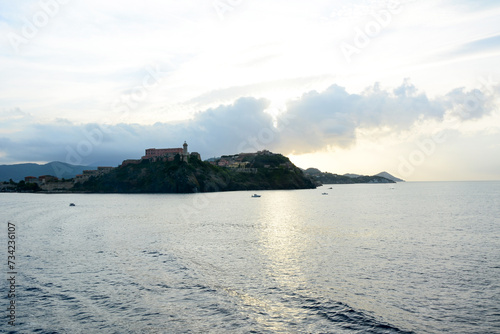 View of an evening seascape with an island and mountains in the clouds behind it. A lighthouse and houses on the shore