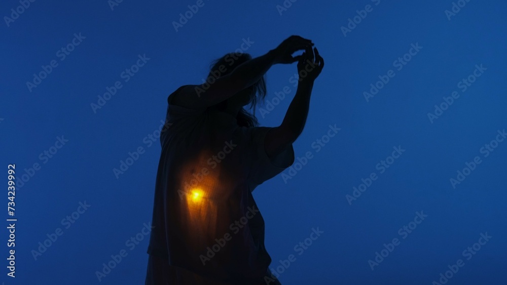 © kinomaster - Female dancer performing in the studio. Young woman dancing in dark studio with warm light glowing inside the chest under her shirt.