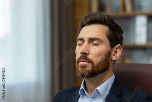 Close-up photo of a young businessman man in a suit sitting in the office at the workplace with closed eyes and resting, seriously thinking.