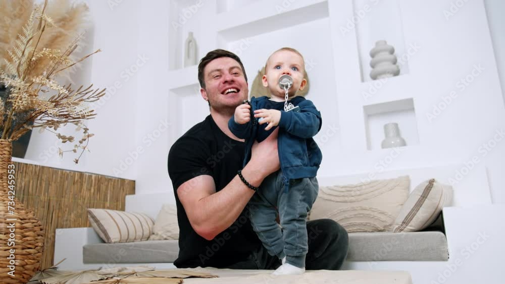 Caucasian male holds an infant child standing on the coffee table. Cute kid with pacifier smiles adorably. Low angle view. Caucasian male holds an infant child standing on the coffee table. Cute kid with pacifier smiles adorably. Low angle view.